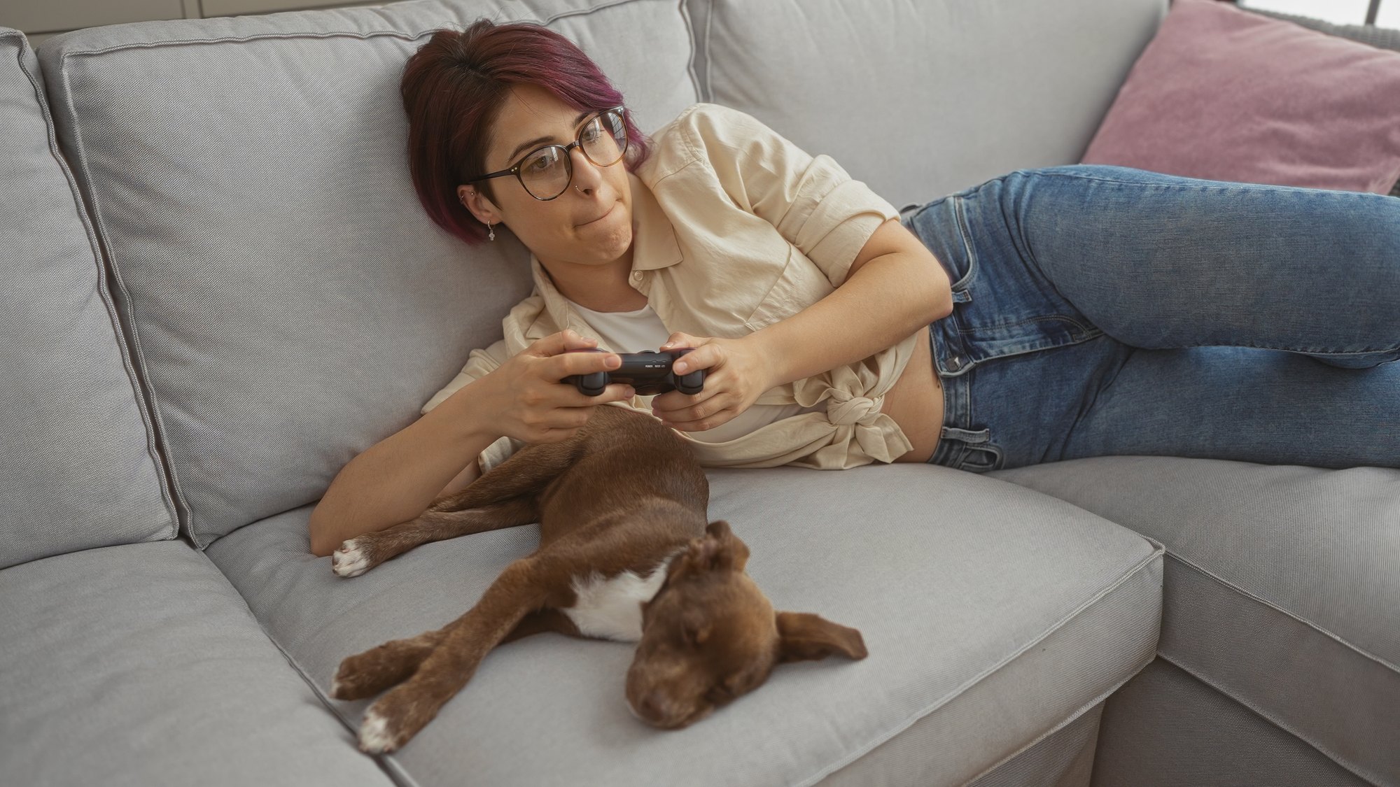 Young woman playing video games while relaxing on a living room sofa with her dog nearby