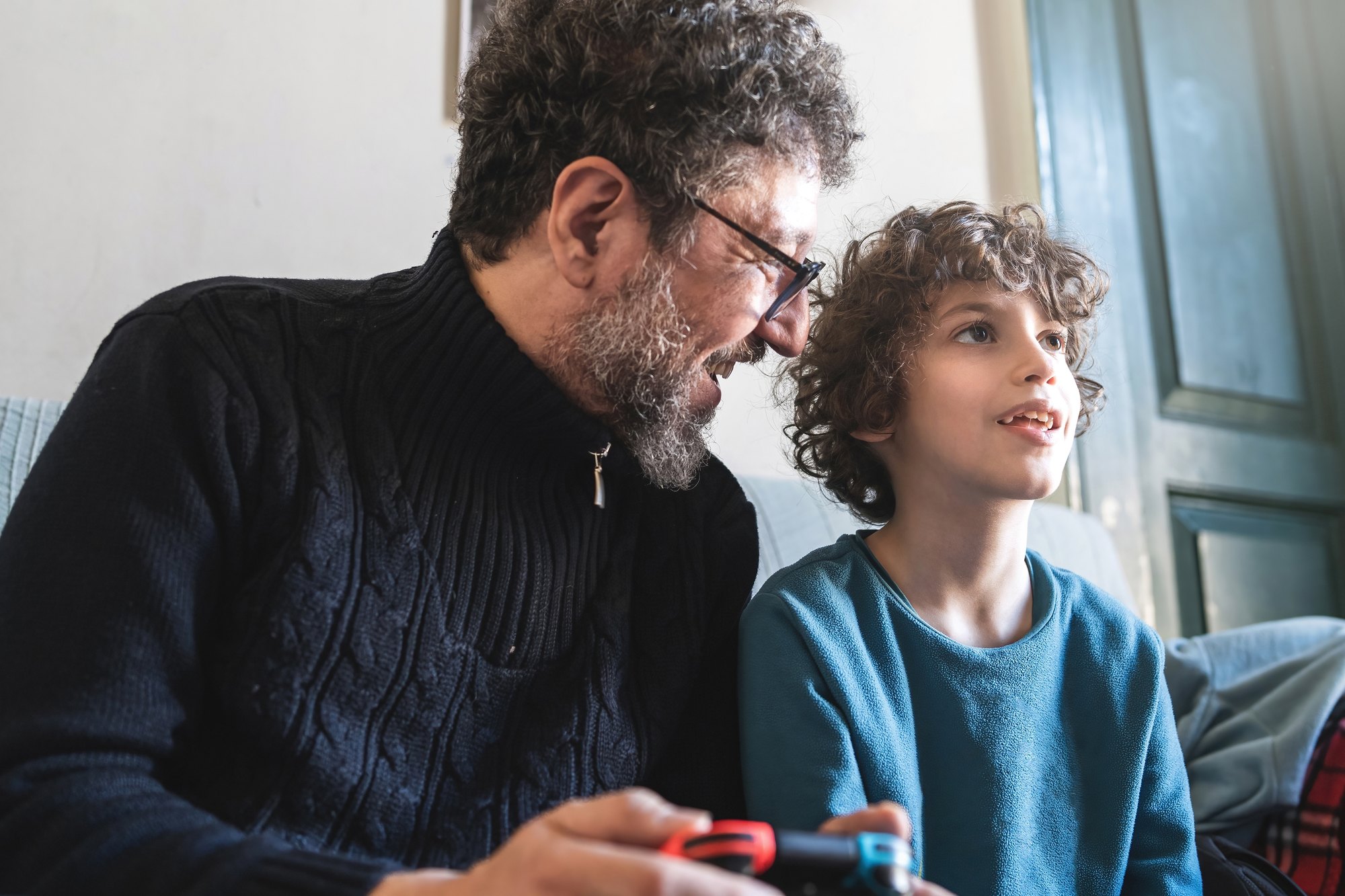 Father and son sitting on the sofa playing videogames together and holding joypad